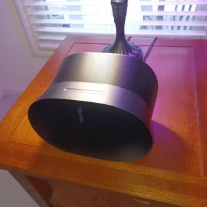Sonos Era 300 smart speaker resting on a wooden side table, illuminated by subtle purple light, with a window in the background for Atmos Clarity